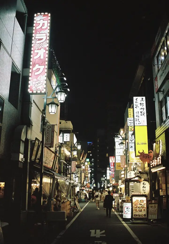 A narrow Tokyo street at night, vertical signs and lanterns, a few people walking away.